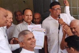 Group of men in white shirts gathered for a microphone speech; one man holds papers and smiles in the foreground.