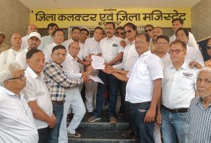 Group of men in white shirts stand on steps, exchanging a document in front of a yellow signboard with Hindi text outside a building.