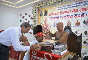 Monk blessing a man wearing a red headband at a ceremony, with a table of books and a microphone nearby.