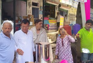 People stand at a busy street-side beverage stall; a woman drinks from a cup while a man in a neon green shirt points, with a large metal urn and shop signs in the background.