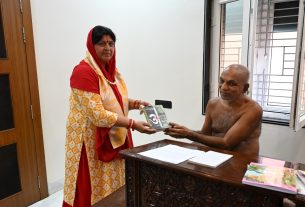 Woman in a red-scarf and patterned orange dress hands a book to a shirtless man seated at a carved wooden desk in a bright room.