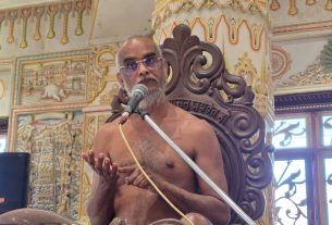 A shirtless man with glasses speaks into a microphone from an ornate carved chair in a richly decorated temple setting.