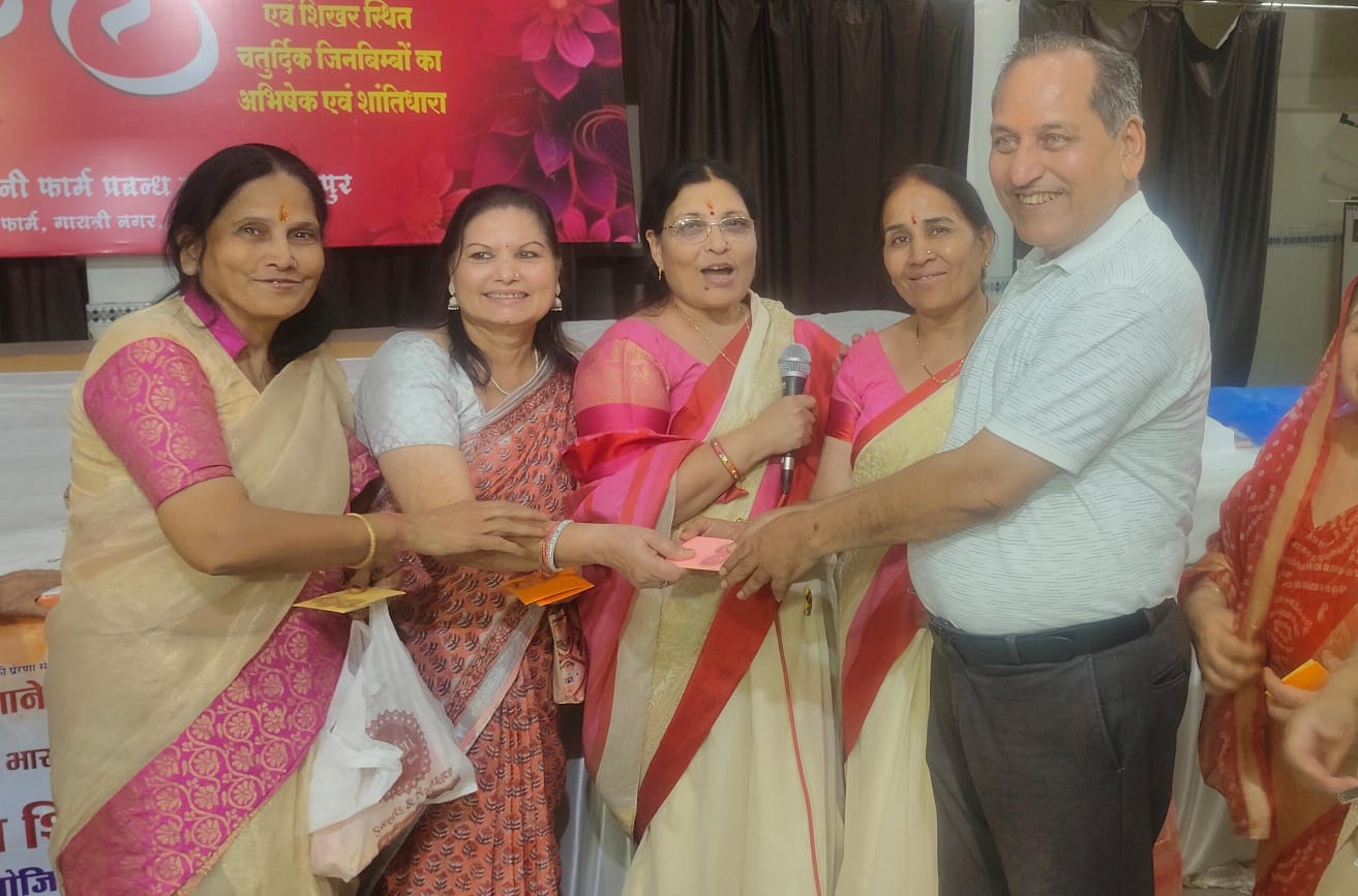 Group of five adults at a cultural/event ceremony; a woman with a microphone hands a pink card to a smiling man while others look on.