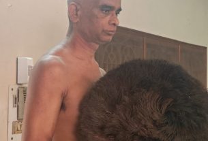Side-profile of a shirtless man with a short haircut indoors, with a large brown fluffy prop obscuring his torso.