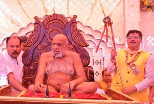 Bearded spiritual leader seated cross‑legged on an ornate throne at a religious ceremony, flanked by two attendants, one holding a ceremonial staff.