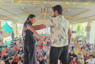 Two adults dance on a stage or open area as a large group of children sits and watches under a covered outdoor space.