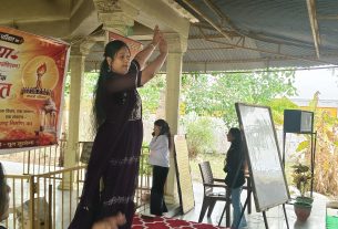 Woman in a dark outfit raises her arms in a yoga pose on a stage at a community event, with large Hindi banners in the background and people watching.