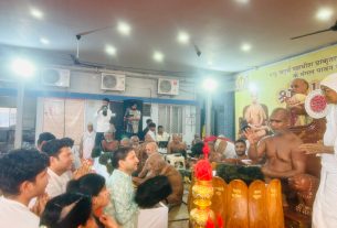Crowd gathered in a temple hall for a ceremony, priests in traditional attire around a brass lamp altar, banner in the background.