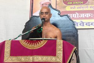 Bald man giving a speech at a podium draped in bright magenta fabric, microphone in front, coconuts lined on the stage, with colorful Hindi posters in the background.