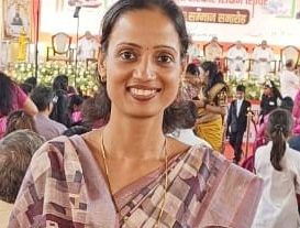 Smiling woman in a patterned saree at an awards ceremony, with a crest badge on her chest and a crowd and banner behind her.