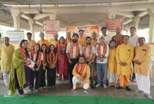 Group of men and women posing for a photo at a community event with Hindi banners in the background on a stage.