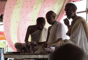 Three monks in white robes gathered around a baby on a decorated table inside a pink-draped tent.
