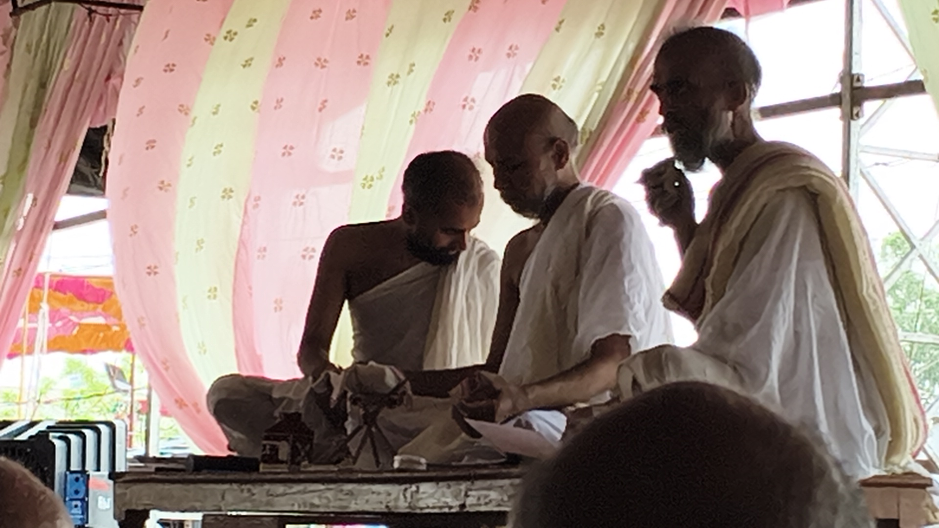 Three monks in white robes gathered around a baby on a decorated table inside a pink-draped tent.