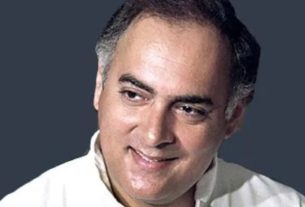 Portrait of a smiling man with short dark hair and a light shirt against a dark gray background.