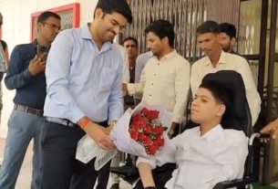 Man in a blue shirt hands a bouquet of red flowers to a woman in a wheelchair, while colleagues stand nearby in a hallway.