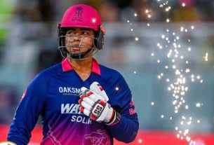 Cricketer in a blue-pink uniform and pink helmet, wearing batting gloves and looking to the side during a celebration with spark effects in the background