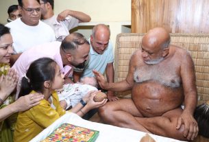 Family members gather around a shirtless elderly man with a white beard, blessing a baby held toward him.
