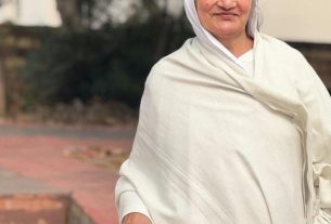 Older woman with gray hair in a white shawl, using a silver cane outdoors on a brick path.