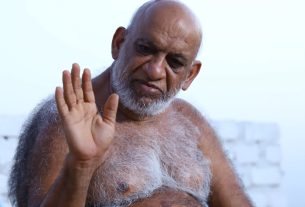 Older man with a white beard waves hello outdoors on a sunny day.