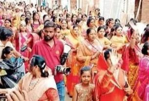 Crowded street festival with people in colorful sarees and traditional dress, some taking photos and waving.
