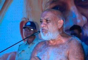 Elderly, bearded man shirtless and speaking into a microphone at a podium during a public event, with blue lighting and a backdrop featuring a large image; a uniformed man sits in the background.
