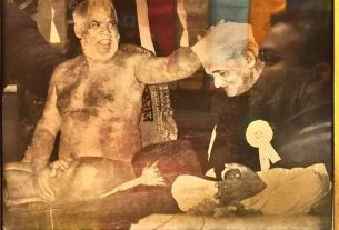 Sepia-toned framed photo of two life-size statues: a nude, muscular man on the left and an older man in a suit with a white rosette on his lap, seated and facing him.