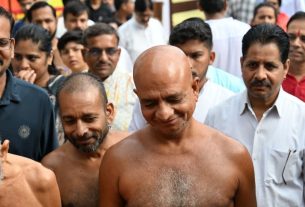 A group of men outdoors, several shirtless with shaved heads, standing close together as onlookers gather behind them.