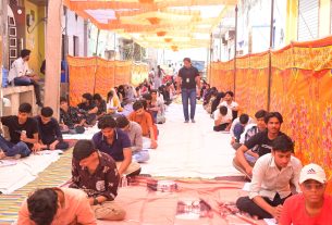 Long line of students sitting on mats and papers, writing in an outdoor tented street covered with orange banners and decorations.