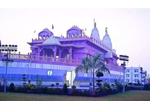 Large ornate temple-like building with domes and spires, set in a landscaped compound with palm trees and outdoor floodlights.