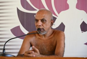 Shirtless man with a gray beard speaking into a microphone at a wooden podium against an abstract purple backdrop