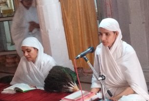 Two people in white traditional attire and turbans sit at a table with open books, one writing and the other speaking into a microphone.