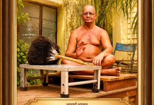 Portrait of a bald, shirtless monk seated cross‑legged on a wooden platform outdoors, with a large feather brush, framed by an ornate gold frame and Hindi plaque.