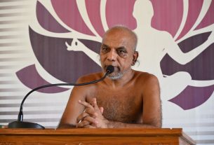 Older man with a gray beard speaks into a microphone at a wooden podium, shirtless, with a purple abstract backdrop behind him.