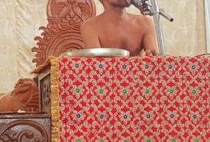 Bald shirtless man speaking into a microphone at a podium draped with a red patterned cloth, ornate wooden backdrop visible behind him.