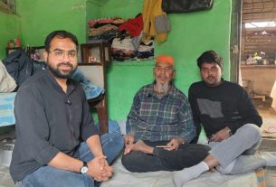 Three men sit cross-legged on a bed in a green room, with clothes and a black bag hanging on the wall behind them.