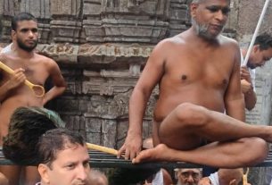 Shirtless man sits cross‑legged on a metal frame during a Hindu temple ritual, surrounded by devotees holding rods, with carved stone temple backdrop behind.