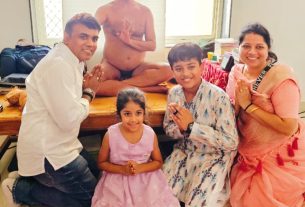 Family group kneeling in prayer around a smiling boy, girl, and woman, with a shirtless man meditating on a table behind them.
