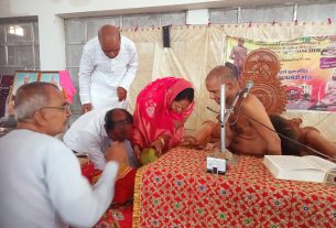 A Hindu ceremony with a shirtless monk at a microphone, villagers bowing beside a richly patterned cloth-covered table. Balance of tradition and prayer scene in a hall.