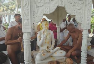 Worshipers surround a seated white marble statue under an ornate canopy during a ritual.
