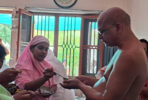 A shirtless man hands a spoonful of food to a woman wearing a pink headscarf at a dining table inside a room, with others nearby.
