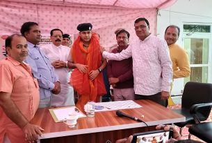 Group of men in traditional Indian attire stand around a wooden table during a ceremony; one man in orange robes with a black beret appears to be the focal figure, while a microphone and papers sit on the table.