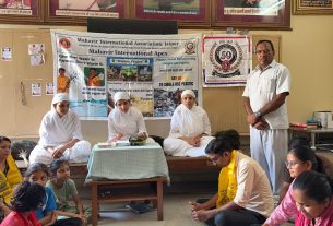 Indoor community gathering: three women in white robes sit at a table in front of banners about environment; a man stands to the right while children listen.