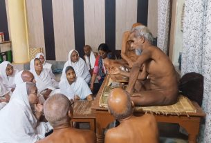 Elderly devotees in white robes sit cross-legged on the floor, watching a ritual performed by barefoot priests on a wooden platform.
