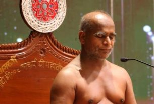 Shirtless man meditating on a carved wooden throne on a stage, with a decorative round plate behind him and a microphone nearby.