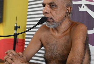 Older, shirtless man with gray beard sits at a table speaking into a microphone, hands clasped, backdrop with yellow wall and striped/purple banner