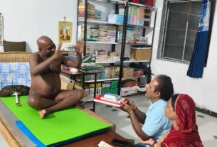 Man sitting cross‑legged on a green mat on a low table, raising his hands in a gesture, as two visitors present a stack of books in a room lined with bookshelves and boxes on top; window with blue curtain and an wall-mounted air conditioner visible.