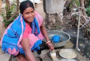 Woman in a pink and blue sari crouches by an outdoor water tap, washing dishes in metal basins amid a rural setting.